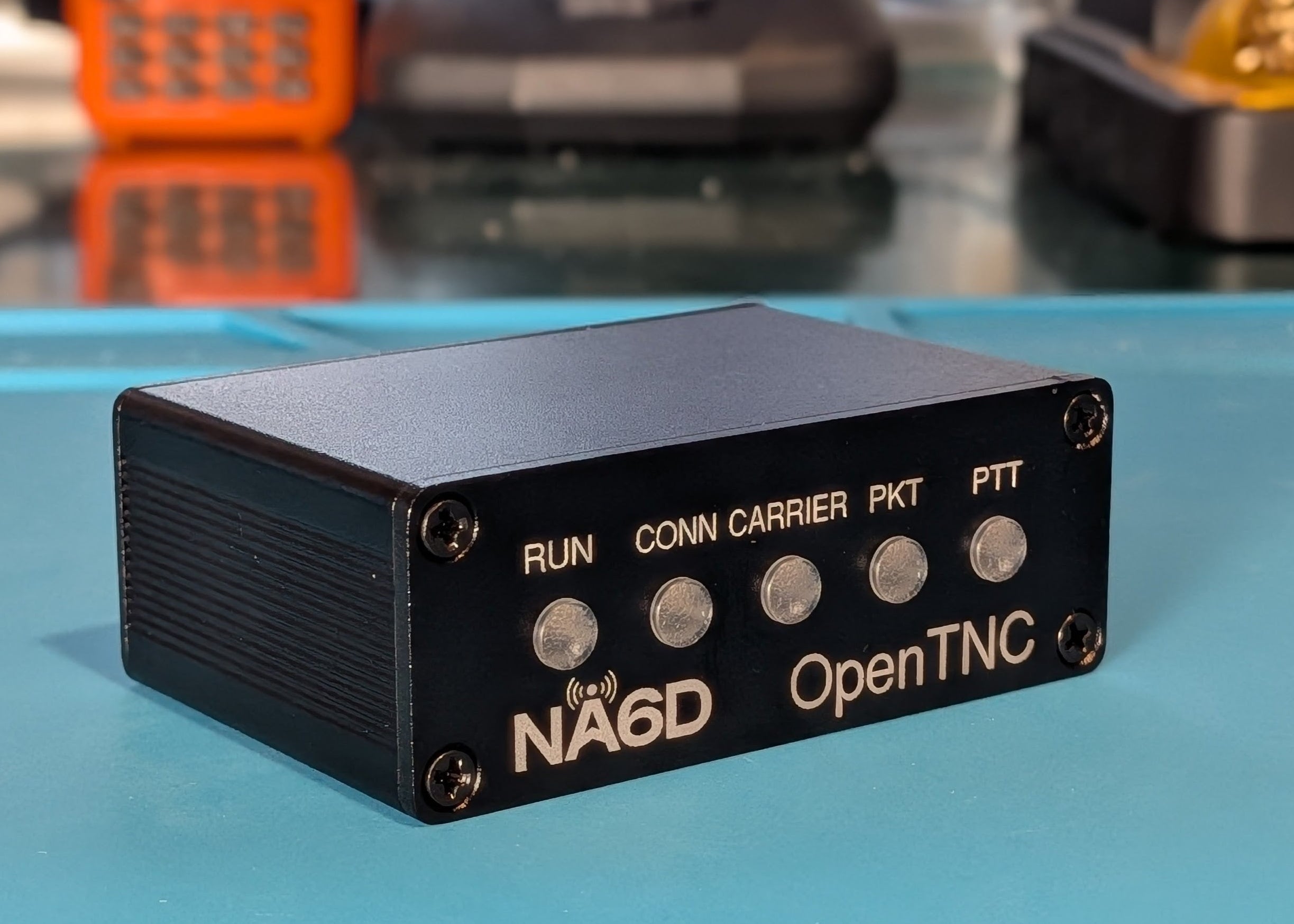 A black metal electronic device labeled 'NA6D OpenTNC' sits on a bright blue surface, with a blurred background showing other electronic equipment.